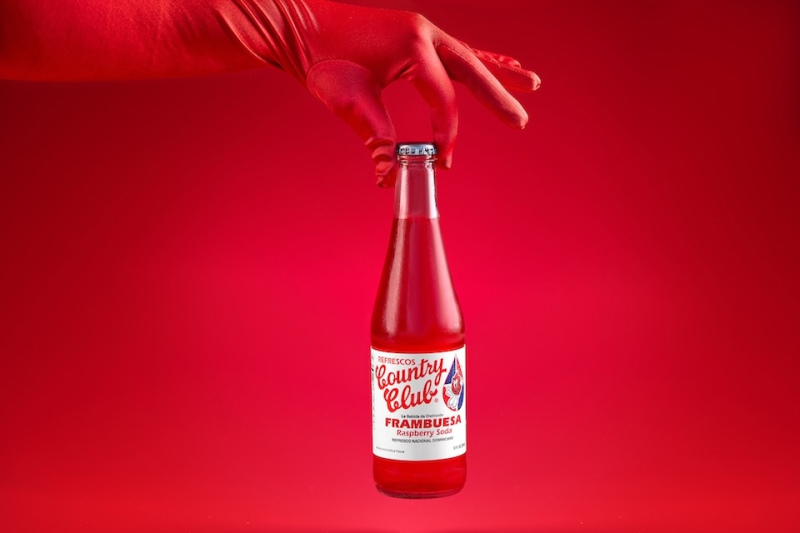 Food photography of a bottled soft drink against a red background  being held by a red-gloved hand