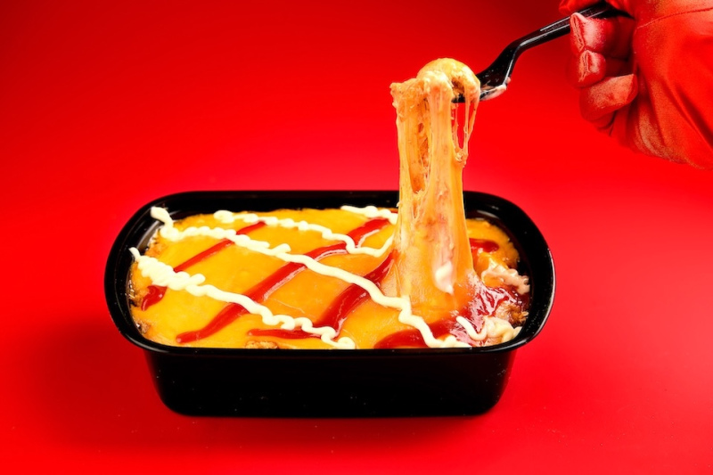 Food photography of a cheese casserole dish on a red background with a red gloved hand doing a cheese pull with a fork