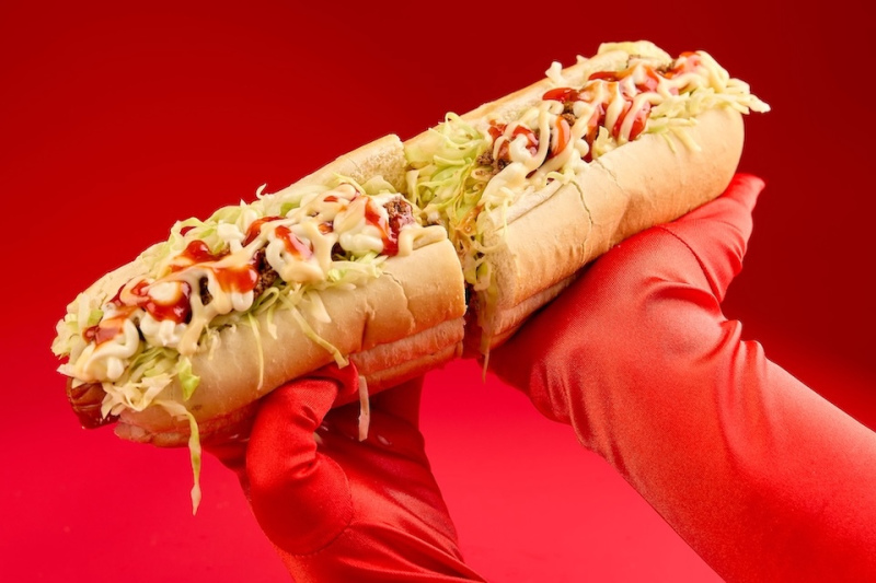 Food photography of a large hot dog loaded with toppings being held up against a red backdrop by two red gloved hands