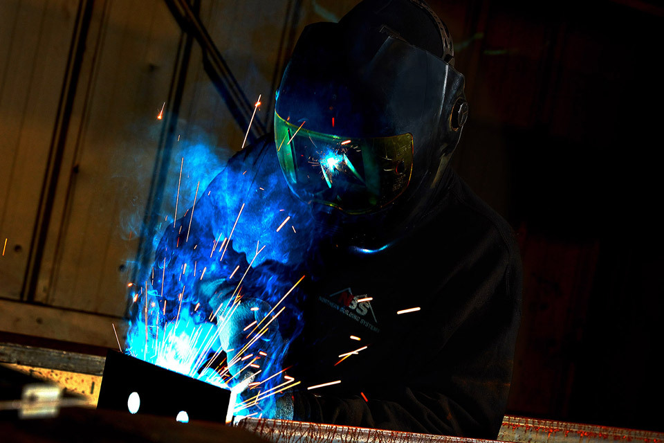 lifestyle photography of a welder with a helmet on welding with blue light and sparks for Package Steel of Sutton, MA.