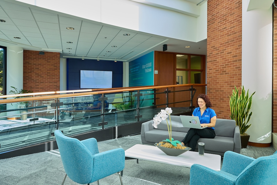 Lifestyle photography of female employee seated on a couch in a hallway typing on a laptop at Teradyne of North Reading, MA.